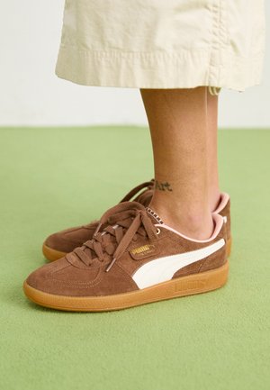 Baskets en daim marron avec accent blanc, semelle en caoutchouc et talon rose. Lacets et détail de marque visibles. Tatouage à la cheville partiellement visible.
