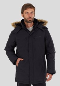 Man in een zwarte winterparka met een capuchon en bontafwerking, met de handen in de zakken, die iets opzij kijkt tegen een effen achtergrond.