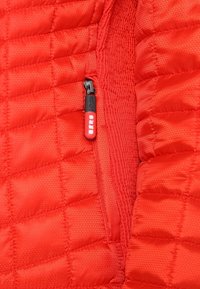 Veste matelassée rouge avec une tirette de fermeture éclair verticale noire et rouge, partiellement dézippée montrant des détails de tissu cousu et rembourré.