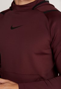 Bordeaux atletisk hoodie med høj krave, sorte accenter og Nike logo. Glat tekstur med tætsiddende design og ribbet detaljer på skuldrene.
