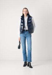 Gilet imbottito blu navy con colletto alto, indossato sopra una camicia bianca; abbinato a jeans azzurri chiari e stivali neri con tacco, mentre tiene una piccola borsa nera.