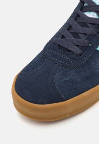 Sapatilha em camurça azul-marinho com um cabedal texturizado, bico redondo e sola de borracha colorida contrastante. Apresenta atacadores escuros e detalhes de costura.