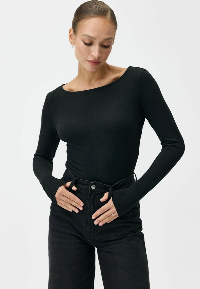 Kobieta w dopasowanej czarnej bluzce z długim rękawem i czarnych dżinsach z wysokim stanem, ręce spoczywają blisko pasa, patrzy w dół na jednolitym tle.