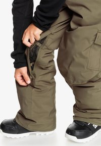 Pantalons de neige kaki avec une ouverture latérale zippée ; tissu texturé ; associés à des bottes de neige noires avec des semelles blanches et un design robuste.
