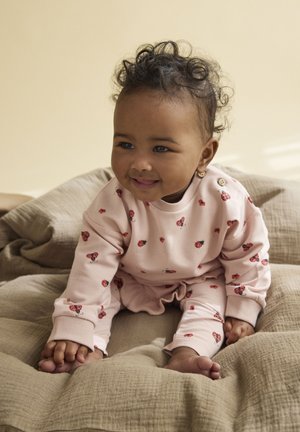 Tenue en coton rose clair avec motifs de coccinelles, comprenant des manches longues et une taille élastique. Enfant assis sur un coussin beige texturé.