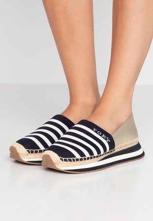 Füße, die schwarz-weiß gestreifte Slip-On-Espadrilles mit beigem Absatz und einem Detail aus gewebtem Seil an der Sohle tragen.
