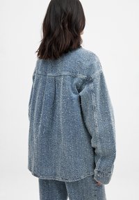 Denim jacket in light blue with a textured, woven finish. Features a relaxed fit, dropped shoulders, and pleated back detail.