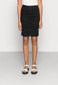 Jupe crayon en denim noir avec une silhouette ajustée, des poches avant et un ourlet discret. Portée avec des sandales blanches à semelles noires.