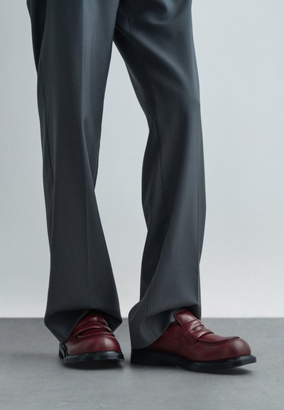 Osoba v voľných šedých nohaviciach a tmavočervených kožených mokasínach stojí na šedom podlahe proti jednoduchému pozadiu.