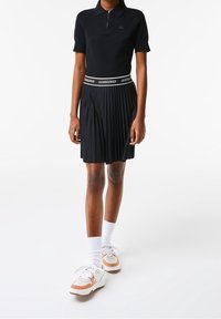 Černé polo s páskem na zip, černá plisovaná sukně s bílým pasem s nápisem "LACOSTE", doplněná bílými a hnědými teniskami.