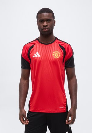 Homme debout, faisant face à l’avant, portant un maillot de sport rouge et noir Manchester United Adidas et un short noir sur un fond uni.