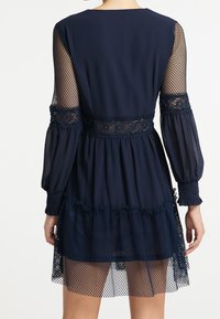 Robe bleu marine avec manches longues en maille transparente, ceinture en dentelle fleurie, et superposition de filet à l'ourlet. Caractéristiques : manches bouffantes et poignets froncés.