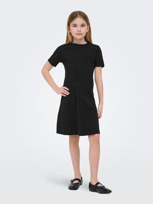 Pige stående med hånden på hoften iført en knælængde sort kortærmet kjole og sorte Mary Jane-sko på en hvid baggrund.