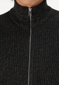 Schwarzer hochgeschlossener Pullover aus Strick mit einem Reißverschluss vorne und strukturiertem Stoff. Die Nähte verleihen dem Design Tiefe.