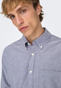 Lyseblå skjorte med knapper og krave, med en brystlomme foran og tekstureret stof. Knapperne er hvide og tydelige.