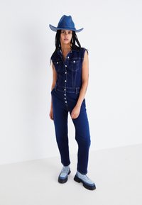 Salopette en denim à design sans manches, lavage bleu foncé, boutons sur le devant et poches à rabat ; associée à un chapeau en denim bleu assorti et des bottes imposantes.