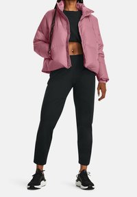 Lichtroze pufferjack met een rits aan de voorkant, voorzien van een hoge kraag; gecombineerd met zwarte tapering broek en zwarte sportschoenen.