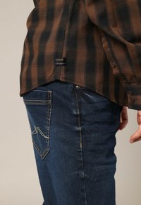 Bruine en zwarte geruite blouse met lange mouwen, gedragen met blauwe jeans. De blouse heeft een label aan de zoom en knopen langs de manchetten.