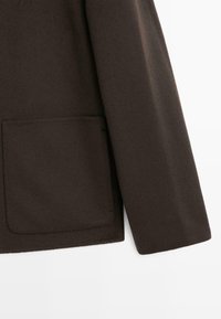 Cappotto marrone in lana con una finitura testurizzata, dotato di una grande tasca frontale e maniche a taglio dritto. Design semplice e minimalista.