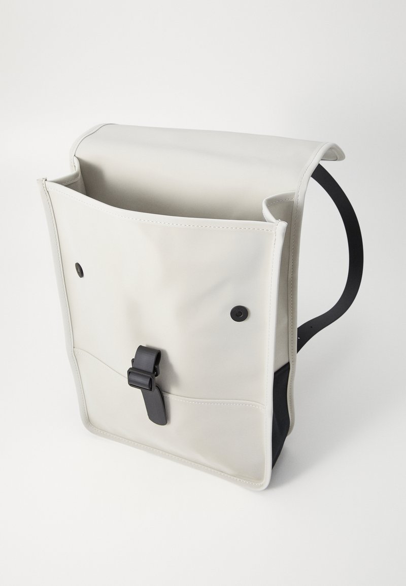 Sac à dos avec un extérieur beige crémeux, doux et lisse, comportant un rabat supérieur, une fermeture à sangle noire et des accents latéraux noirs.