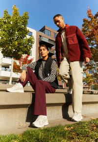 Černá vesta přes pruhovaný top; vínové kalhoty; krémové tenisky. Muž v vínové fleecové bundě s kapsou, světlé kalhoty a krémové tenisky.