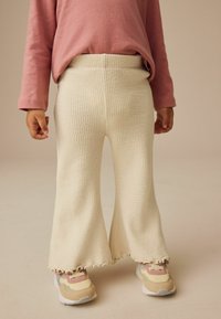 Pantaloni flare a maglia color crema con un motivo testurizzato e orlo a scallop, abbinati a sneakers con accenti pastello e una maglietta a maniche lunghe rosa.