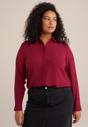 Maroon long-sleeve blouse with a button-up collar, smooth texture, relaxed fit, and a curved hem, paired with a black denim skirt.
