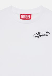 Biely bavlnený tričko s okrúhlym výstrihom a čierne vyšívané logo "Diesel" na hrudi. Červený štítok značky na golieri.