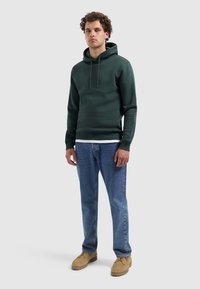 Donkergroene hoodie met een voorzak, verstelbare capuchon en geribde mouwen, gecombineerd met lichtblauwe denim jeans en beige moccasin schoenen.