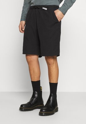 Homme portant un short noir mi-genoux avec une ceinture, des chaussettes noires et des bottines en cuir noir, debout devant un fond uni.