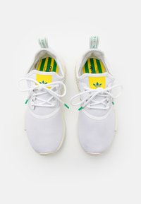 Zapatillas deportivas blancas con material de malla, punta redonda y acentos decorativos en amarillo y verde en la lengüeta y las plantillas. Incluyen cordones planos y una tira de agarre.