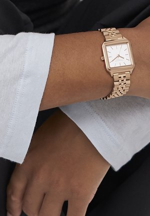 Rechteckige roségoldene Armbanduhr mit einem Gliederarmband an einer Person, die ein hellblaues Langarmhemd trägt.