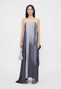 Grijze asymmetrische maxi-jurk met een soepele, zijdeachtige textuur, voorzien van dunne bandjes en een subtiel verticaal streepjespatroon. Inclusief kleine zilveren handtas.