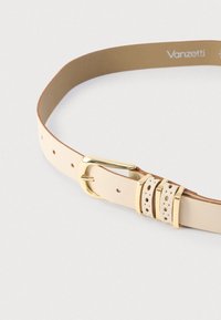 Beigen värinen nahkavyö, jossa on kullanvärinen metallisolki. Vyössä on sileä pinta ja kaksi riviä pieniä reikiä remmissä säätömahdollisuutta varten.