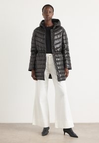 Doudoune noire avec capuche, fermeture éclair et cordon de serrage élastique. Présente un extérieur brillant et une silhouette près du corps. Associée à un pantalon ample blanc.