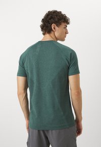 Hombre con cabello rizado que lleva una camisa corta de manga verde texturizada y pantalones cortos grises, visto desde atrás contra un fondo blanco liso.