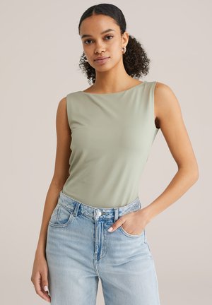 Göndör hajú nő világoszöld ujjatlan felsőben és világoskék farmerben, zsebre dugott kézzel pózol bézs háttér előtt.