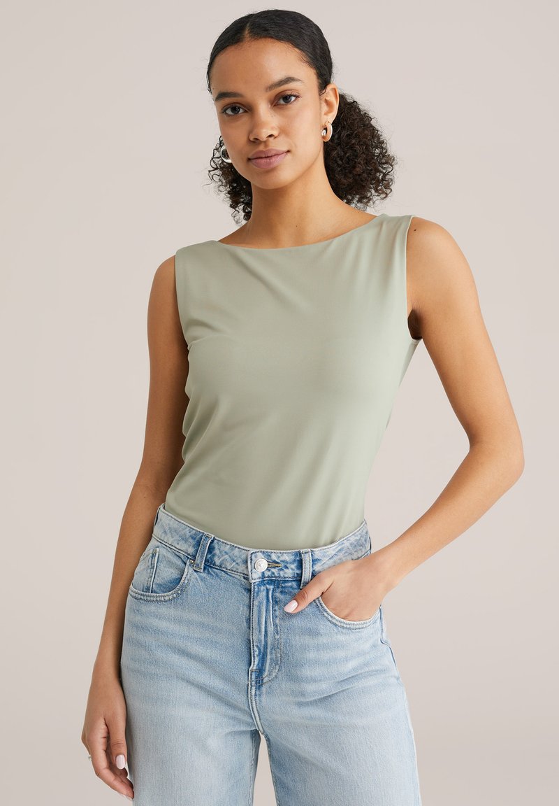 Göndör hajú nő világoszöld ujjatlan felsőben és világoskék farmerben, zsebre dugott kézzel pózol bézs háttér előtt.