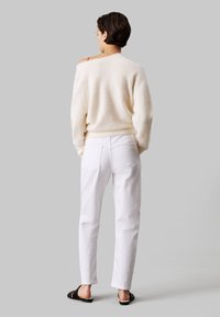 Białe spodnie o luźnym kroju w połączeniu z puszystym kremowym swetrem; sweter ma fason odkrywający ramiona. Na stopach czarne klapki.