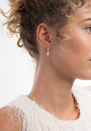 Boucles d'oreilles créoles en or avec un pendentif circulaire en pierre, présentant une surface texturée, portées par une personne aux cheveux bouclés.