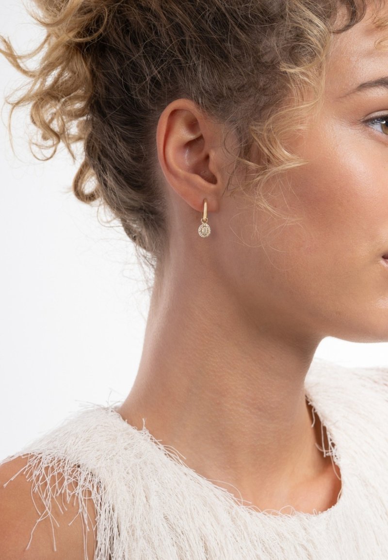 Boucles d'oreilles créoles en or avec un pendentif circulaire en pierre, présentant une surface texturée, portées par une personne aux cheveux bouclés.