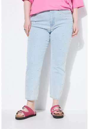 Hellblaue Jeans mit kleinen weißen Perlenakzenten, kombiniert mit pinkfarbenen Sandalen mit offenen Zehen und einem pinkfarbenen Hemd, stehend vor weißem Hintergrund.