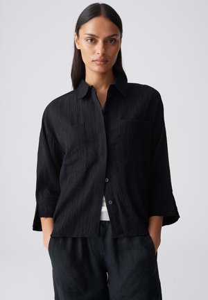 Donna con lunghi capelli scuri indossa una camicia nera texturizzata con bottoni e pantaloni neri, in piedi con le mani nelle tasche davanti a uno sfondo semplice.