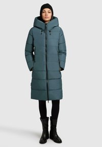 Teal polstret lang jakke med høj krave, frontlynlås og to sidelommer. Båret over sorte leggings og ankelstøvler, hvilket skaber et lagdelt look.