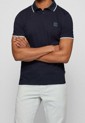 Muž se zkříženýma rukama, oblečený v tmavě modrém polotriku s bílým lemováním a bílých kalhotách, na jednobarevném pozadí.