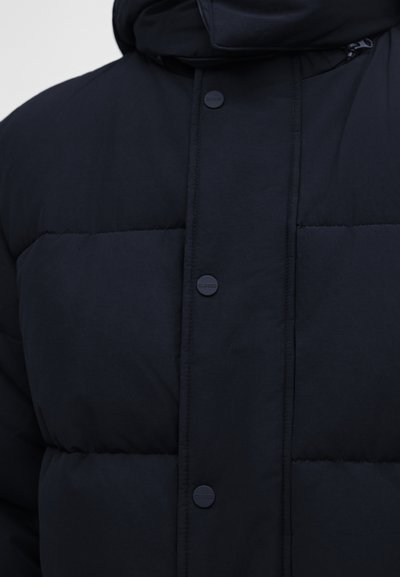 Gros plan d'une veste matelassée noire avec boutons-pression et coutures visibles à l'avant ainsi qu'un col haut.
