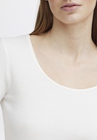 Hvit ribbet topp med rund hals, tettsittende passform og teksturert stoff. Fokus ligger på plaggets øvre del og halsåpning.