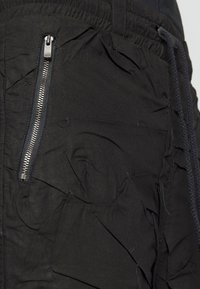 Pantalon en tissu noir avec un gros plan sur une poche zippée verticale argentée et des détails de couture texturés près de la taille.