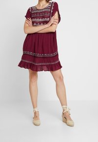 Bordová šatová šaty s krátkým rukávem, vyšívanými detaily, volánkovým lemem a volným střihem. V kombinaci s béžovými espadrilovými wedges.