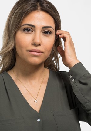 Goldene Halskette mit einem Herzenanhänger, der einen kleinen Kristall enthält. Das Modell trägt eine olivgrüne Bluse mit V-Ausschnitt und geknöpften Ärmeln.